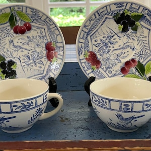 May include: Two white teacups and saucers with blue floral and bird designs. The saucers feature hand-painted berries. The cups and saucers are displayed on a blue surface, creating a charming tea set.
