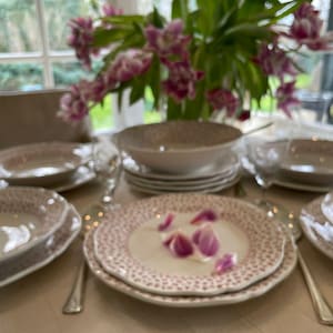 Pode incluir: Uma mesa posta com pratos, tigelas e talheres brancos e rosa com bolinhas. Um buquê de flores rosas e roxas está ao fundo. A configuração está em uma toalha de mesa bege, criando uma atmosfera elegante.