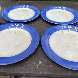 Könnte beinhalten: Vier runde Keramikteller mit einem breiten kobaltblauen Rand und einer cremefarbenen Mitte, die mit einem handgemalten grünen Stängel und einer weißen Blume verziert ist. Zwei klare Weingläser mit blau getönten Stielen stehen an den Seiten.