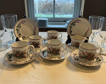 Op de afbeelding: Een theeservies van wit porselein met bloemmotieven en een bordeauxrode rand. De set bestaat uit kopjes, schotels en borden. Heldere wijnglazen staan ernaast. Op een wit tafelkleed.