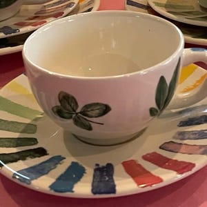 Puede incluir: Un juego de taza de t&eacute; y plato de cer&aacute;mica blanca con un dise&ntilde;o pintado a mano de hojas verdes en la taza y un patr&oacute;n radial colorido de pinceladas cortas y gruesas en amarillo, verde, azul, morado y rojo en el borde del plato.