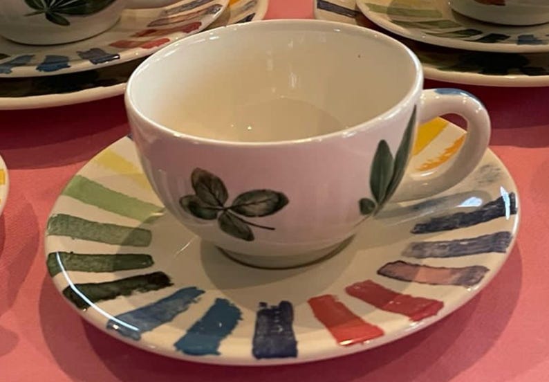 Puede incluir: Un juego de taza de t&eacute; y plato de cer&aacute;mica blanca con un dise&ntilde;o pintado a mano de hojas verdes en la taza y un patr&oacute;n radial colorido de pinceladas cortas y gruesas en amarillo, verde, azul, morado y rojo en el borde del plato.