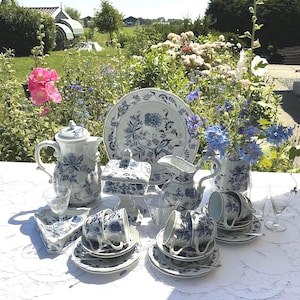 Peut inclure: Un service à thé complet en porcelaine bleue et blanche, comprenant une théière, un sucrier, une crémière, des tasses, des soucoupes et des assiettes de service. L'ensemble présente un motif floral et est disposé sur une nappe blanche.