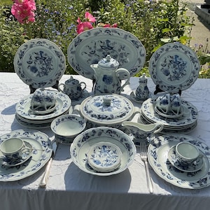 May include: A full blue and white porcelain dinner service, including plates, cups, saucers, bowls, a teapot, serving dishes, and a platter. The set features a floral design and is arranged on a white tablecloth.