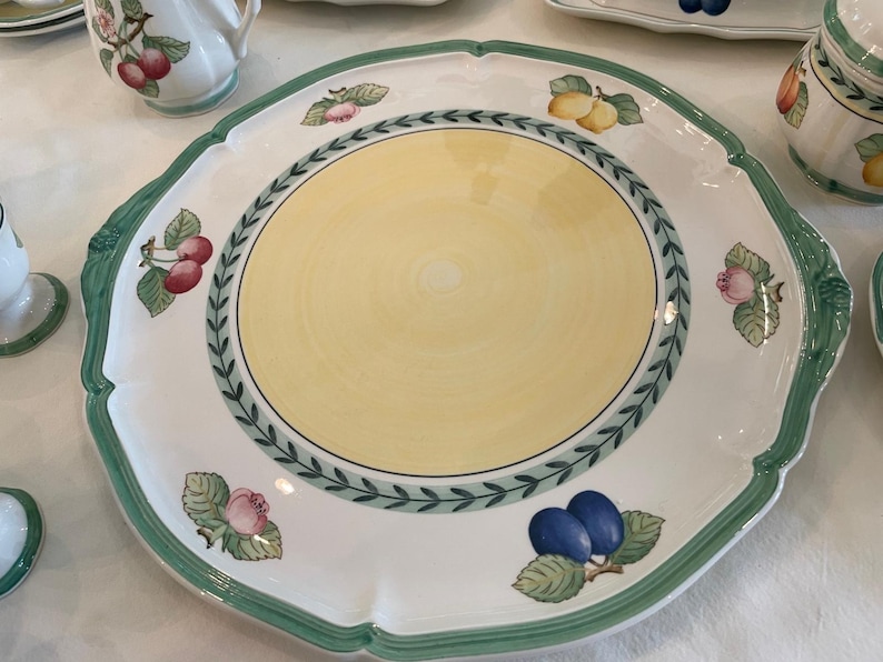 May include: A large round ceramic serving platter with a scalloped edge and a green rim. The centre is pale yellow, surrounded by a green leaf border. The white border features illustrations of cherries, lemons, plums, and flowers.