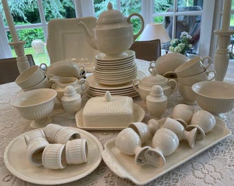 May include: A cream-coloured porcelain dinner service, including a teapot, cups, saucers, bowls, and serving dishes. The set has a ribbed design and is arranged on a white tablecloth, suitable for a formal dining setting.