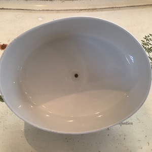 Op de afbeelding: Een witte, ovaalvormige keramische kom met een klein gaatje in het midden. De kom heeft een glanzende afwerking en staat tegen een achtergrond met botanische illustraties. Het ontwerp van de kom is eenvoudig en elegant.