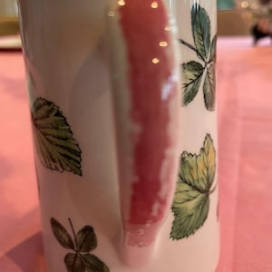 Puede incluir: Una taza de cer&aacute;mica blanca con un patr&oacute;n bot&aacute;nico de hojas verdes e ilustraciones de tr&eacute;boles. La taza tiene un asa vertical prominente con una franja pintada de color rosa rojizo en el centro, que contrasta con el cuerpo blanco del recipiente.