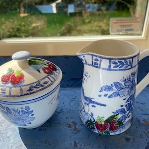 Peut inclure: Un sucrier en céramique avec un couvercle et un petit pichet. Les deux sont blancs avec des motifs floraux et fruités bleus. Le couvercle du sucrier a un bouton bleu et un motif de fraises et de cerises. Le pichet a une anse et un bec.