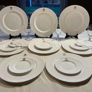 Op de afbeelding: Een verzameling witte porseleinen borden, saladeborden en wijnglazen. Elk bord heeft een gouden wapen. De borden zijn gerangschikt op een wit tafelkleed, sommige op zwarte standaards. Vorken en messen zijn ook zichtbaar.