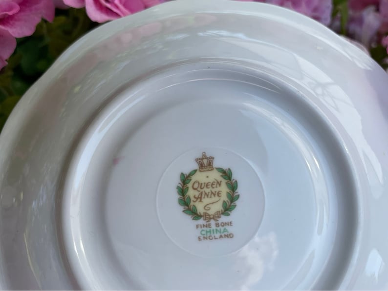 May include: White saucer with a green and gold Queen Anne Fine Bone China England logo in the center.