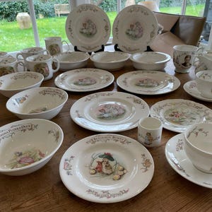 Peut inclure: Ensemble de vaisselle en céramique blanche ornée d'illustrations de personnages de Beatrix Potter. L'ensemble comprend des assiettes, des bols, des tasses, des soucoupes et des mugs, avec des motifs floraux et de personnages aux couleurs pastel.