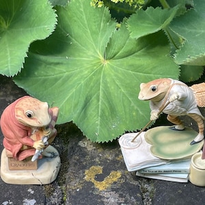 Op de afbeelding: Twee kikkerbeeldjes, een speelt klarinet en de ander houdt een wandelstok vast, zitten op boeken. De kikker die klarinet speelt draagt een rood jasje en de andere kikker draagt een wit jasje. De kikker die klarinet speelt zit op een boek met de tekst "Cream Crackers" erop. De andere kikker zit op een boek met de tekst "The Tale of Mr. Jeremy Fisher" erop.