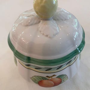 May include: A white ceramic sugar bowl with a lid featuring a yellow fruit-shaped handle. The bowl has a green rim and a decorative band with green leaves and a central illustration of two orange-red fruits with green leaves on the side.