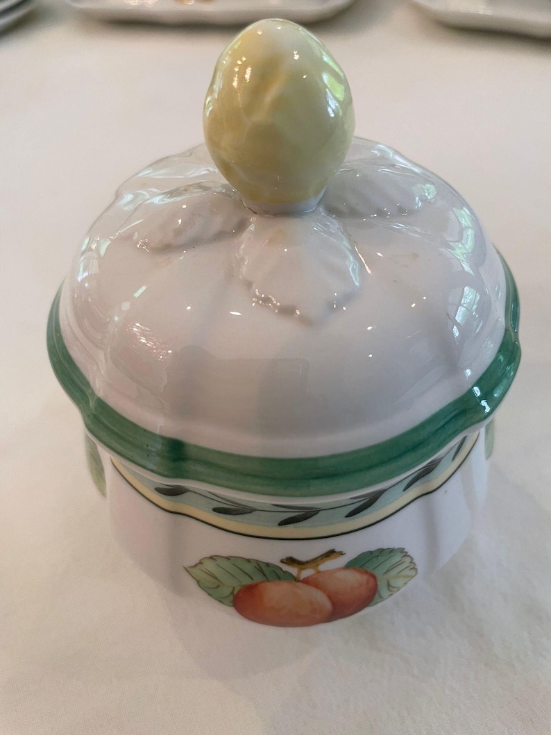 May include: A white ceramic sugar bowl with a lid featuring a yellow fruit-shaped handle. The bowl has a green rim and a decorative band with green leaves and a central illustration of two orange-red fruits with green leaves on the side.