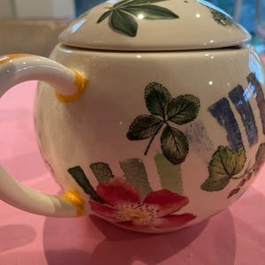 Puede incluir: Una tetera redonda de cer&aacute;mica con tapa, que presenta una base de color crema decorada con hojas verdes pintadas, flores rojas y acentos de pinceladas verticales azules. La tetera tiene un asa curva con detalles pintados en amarillo.
