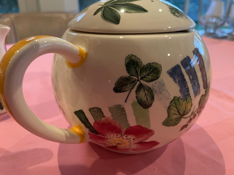 Puede incluir: Una tetera redonda de cer&aacute;mica con tapa, que presenta una base de color crema decorada con hojas verdes pintadas, flores rojas y acentos de pinceladas verticales azules. La tetera tiene un asa curva con detalles pintados en amarillo.