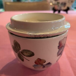 Puede incluir: Un tarro de cer&aacute;mica blanca con tapa que presenta un patr&oacute;n bot&aacute;nico de hojas verdes y flores multicolores. La tapa tiene una muesca visible en el borde exterior. El recipiente tiene una forma redondeada y un acabado suave y brillante.