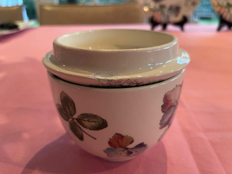 Puede incluir: Un tarro de cer&aacute;mica blanca con tapa que presenta un patr&oacute;n bot&aacute;nico de hojas verdes y flores multicolores. La tapa tiene una muesca visible en el borde exterior. El recipiente tiene una forma redondeada y un acabado suave y brillante.