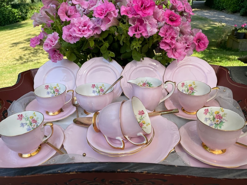 May include: A pink tea set with floral designs on the cups and saucers. The set includes a teapot, creamer, sugar bowl, and six teacups and saucers.