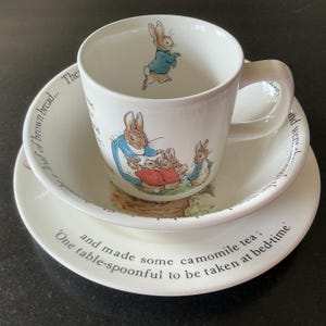 Puede incluir: Un juego de vajilla de tres piezas de cerámica blanca con ilustraciones de Peter Rabbit y texto impreso. El juego incluye una taza con un conejo con chaqueta azul, un cuenco y un plato, decorados con escenas y texto narrativo del cuento.