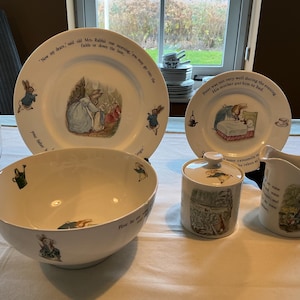May include: A set of white ceramic tableware with illustrations and quotes from a children's story. Includes plates, a bowl, a sugar bowl, and a creamer. The plates feature rabbit illustrations and text around the edge.