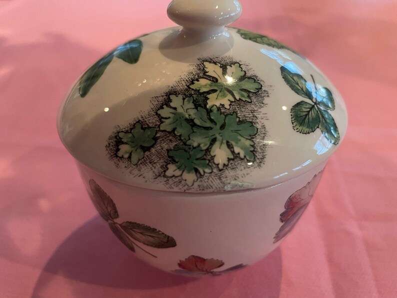 Puede incluir: Un azucarero de cer&aacute;mica blanca con tapa a juego, que presenta un patr&oacute;n bot&aacute;nico de hojas verdes y peque&ntilde;as flores coloridas. El cuenco tiene una forma redondeada y un acabado liso y brillante, con el dise&ntilde;o floral impreso en la superficie.