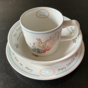 Puede incluir: Un juego de vajilla de tres piezas de cerámica blanca con ilustraciones de Peter Rabbit. Incluye una taza, un cuenco y un plato, decorados con bordes azules, texto en cursiva y las fechas 1893-1993. El interior de la taza muestra el texto Peter Rabbit.