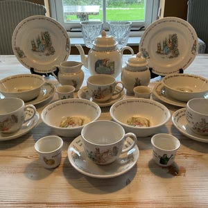 Peut inclure: Un service de table en céramique blanche orné d'illustrations et de textes de Pierre Lapin. L'ensemble comprend deux grandes assiettes, trois tasses avec soucoupes, trois bols, une théière, un pot à lait, un sucrier et trois petits gobelets décorés de scènes colorées.