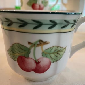 May include: A white ceramic teacup featuring a painted illustration of two red cherries with green leaves on a brown stem. The rim is decorated with a green and yellow leaf-patterned border. The cup has a white handle and a scalloped edge design.