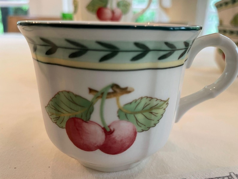 May include: A white ceramic teacup featuring a painted illustration of two red cherries with green leaves on a brown stem. The rim is decorated with a green and yellow leaf-patterned border. The cup has a white handle and a scalloped edge design.
