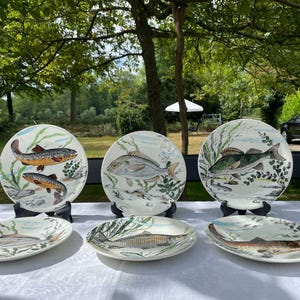 May include: A collection of six ceramic plates with hand-painted fish designs. The plates are in various shapes and sizes, with round and oval options. The fish are depicted in natural colors, with aquatic plants and a light blue background.
