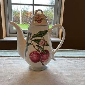 May include: White ceramic teapot with a lid, featuring a handle and spout. The teapot is decorated with a botanical design of peaches, pink flowers, and green leaves. The teapot is sitting on a white tablecloth.