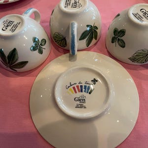 Puede incluir: Tres tazas de t&eacute; de cer&aacute;mica blanca y un platillo de Gien France. Cada taza presenta ilustraciones de hojas verdes y un asa. La base del platillo muestra el texto Couleurs du temps, un logotipo de Gien France y un dise&ntilde;o gr&aacute;fico colorido.