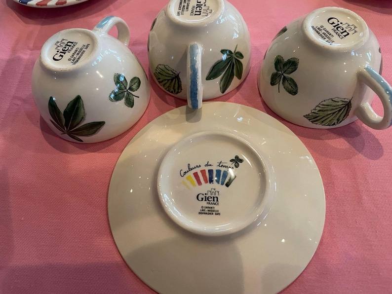 Puede incluir: Tres tazas de t&eacute; de cer&aacute;mica blanca y un platillo de Gien France. Cada taza presenta ilustraciones de hojas verdes y un asa. La base del platillo muestra el texto Couleurs du temps, un logotipo de Gien France y un dise&ntilde;o gr&aacute;fico colorido.