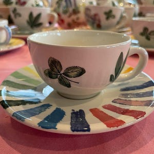 Puede incluir: Un juego de taza y plato de cer&aacute;mica blanca. La taza presenta ilustraciones de hojas verdes en el exterior. El plato est&aacute; decorado con un patr&oacute;n radial de gruesas pinceladas de colores, similares a la pintura, en tonos azul, rojo, amarillo y verde.