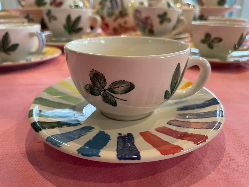 Puede incluir: Un juego de taza y plato de cer&aacute;mica blanca. La taza presenta ilustraciones de hojas verdes en el exterior. El plato est&aacute; decorado con un patr&oacute;n radial de gruesas pinceladas de colores, similares a la pintura, en tonos azul, rojo, amarillo y verde.