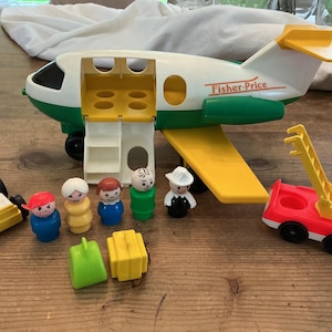 Puede incluir: Un avión de juguete Fisher-Price vintage con un cuerpo blanco y verde, una rampa amarilla y un camión de bomberos rojo. Incluye varias figuras y accesorios de plástico pequeños. El avión tiene el logotipo de Fisher-Price.