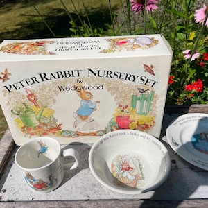 Peut inclure: Un ensemble de puériculture Peter Rabbit par Wedgwood comprenant une tasse, un bol et une assiette en céramique blanche, tous ornés d'illustrations de Peter Rabbit. Les articles sont présentés devant leur boîte en carton blanc d'origine.