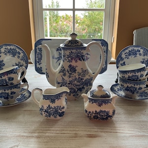 Pode incluir: Um conjunto de chá em cerâmica com base branca e padrão floral azul. O conjunto inclui um bule com tampa, uma leiteira, um açucareiro com tampa, quatro chávenas com pires a condizer e um grande tabuleiro retangular com bordas florais.