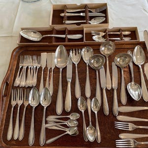 May include: A collection of silver-colored flatware, including forks, spoons, knives, and serving utensils, arranged on a wooden tray. Some pieces are in their original boxes. The flatware has a polished finish, reflecting the light.