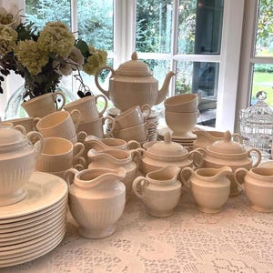 Könnte beinhalten: Eine Sammlung cremefarbener Keramikgeschirr, darunter eine Teekanne, Tassen, Untertassen, Zuckerdosen, Sahnekännchen und Servierschalen. Das Set ist auf einer Spitzendecke angeordnet, mit einem Blumengesteck im Hintergrund.