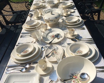May include: A white tablecloth with a lace pattern is set with a formal dinner setting. The table is decorated with white plates with floral designs, silver cutlery, and glasses. A large white serving bowl with a floral design is in the center of the table.