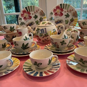 Puede incluir: Un juego de t&eacute; de cer&aacute;mica que incluye una tetera, una lechera, un azucarero y varias tazas con sus platos. Cada pieza est&aacute; decorada con motivos bot&aacute;nicos pintados a mano, con flores rojas y hojas verdes, junto a bordes coloridos con rayas tipo pincelada.