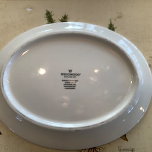 Op de afbeelding: Witte ovale serveerschaal met een licht verhoogde rand. De onderkant toont het Wedgwood-logo en de tekst "SARAH'S GARDEN ENGLAND 2004". De schaal is waarschijnlijk van porselein en geschikt voor het serveren van voedsel.