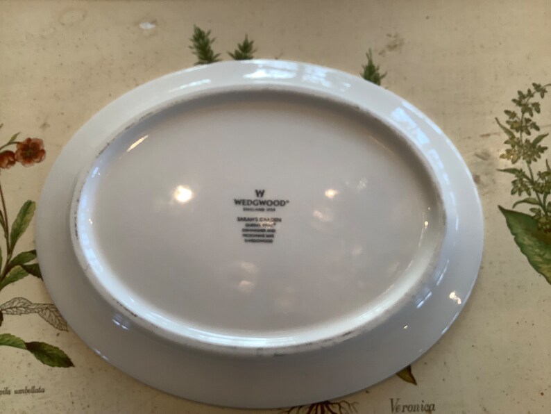 Op de afbeelding: Witte ovale serveerschaal met een licht verhoogde rand. De onderkant toont het Wedgwood-logo en de tekst "SARAH'S GARDEN ENGLAND 2004". De schaal is waarschijnlijk van porselein en geschikt voor het serveren van voedsel.