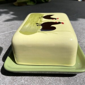 Peut inclure: Un beurrier en c&eacute;ramique vert avec un couvercle jaune orn&eacute; d'un motif peint de deux poulets noirs &agrave; cr&ecirc;tes rouges.