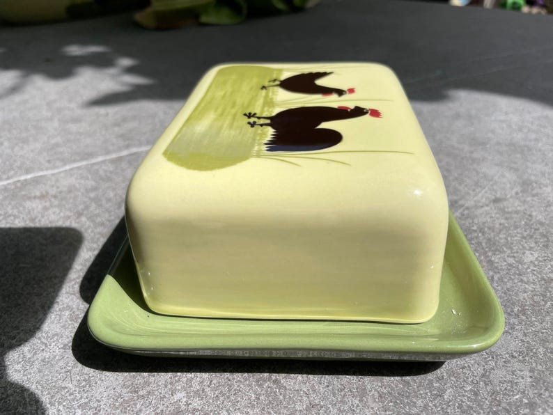 Peut inclure: Un beurrier en c&eacute;ramique vert avec un couvercle jaune orn&eacute; d'un motif peint de deux poulets noirs &agrave; cr&ecirc;tes rouges.