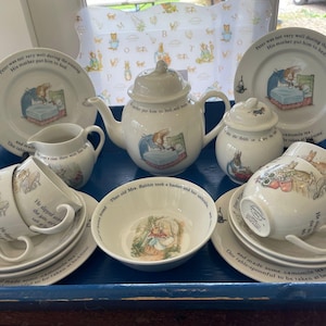 Può includere: Servizio da tè in ceramica bianca con illustrazioni di un coniglio con cappotto blu. Il set include piatti, tazze, piattini, una teiera, una lattiera, una zuccheriera e una ciotola. Testo visibile su piatti e tazze.