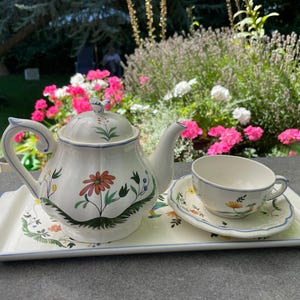 May include: A white ceramic teapot and teacup with floral designs on a white ceramic tray. The teapot has a blue handle and spout, and the teacup has a blue handle. The tray has a blue border.